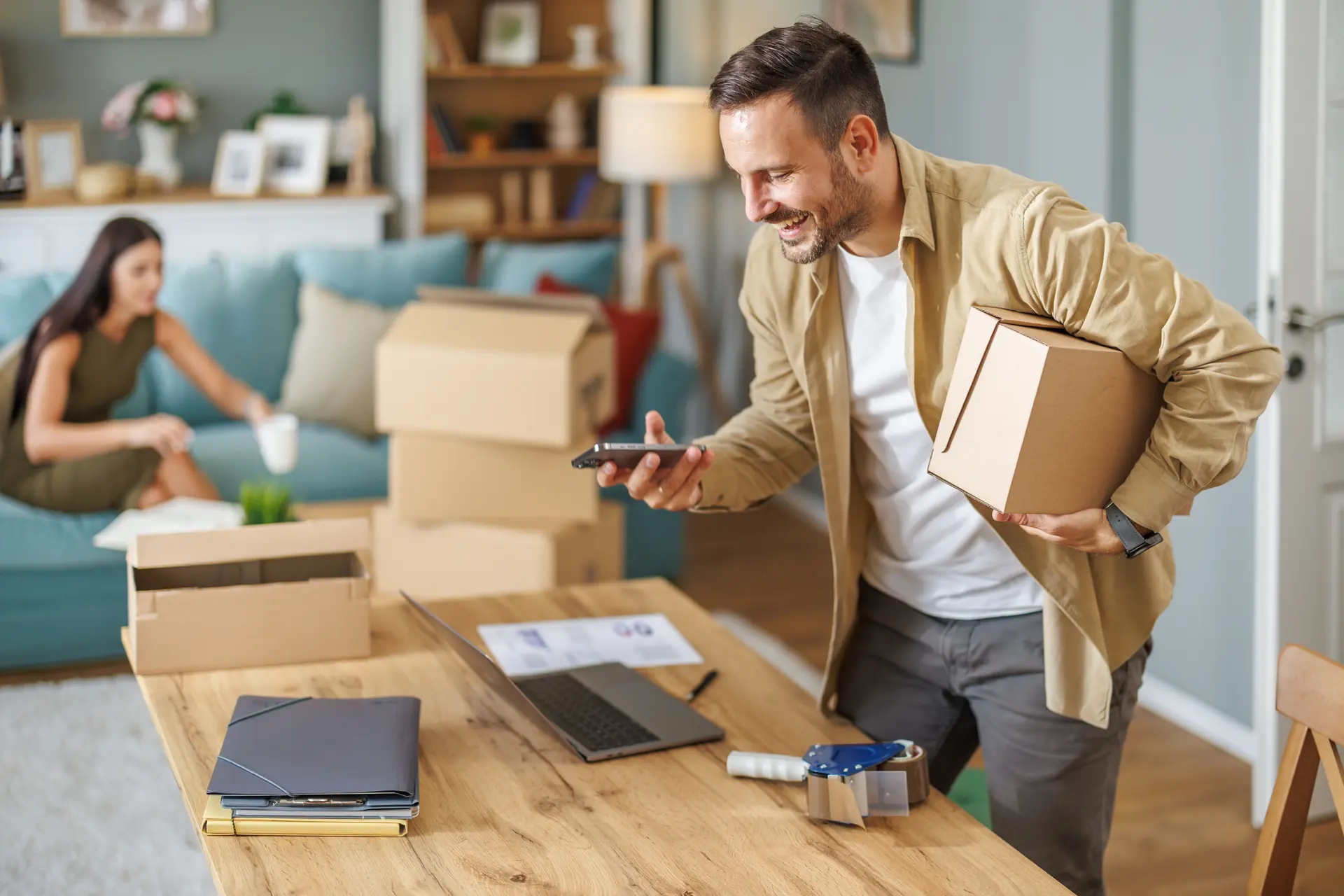 Entrepreneur souriant portant une boîte d’expédition tout en consultant son téléphone dans son espace de travail.